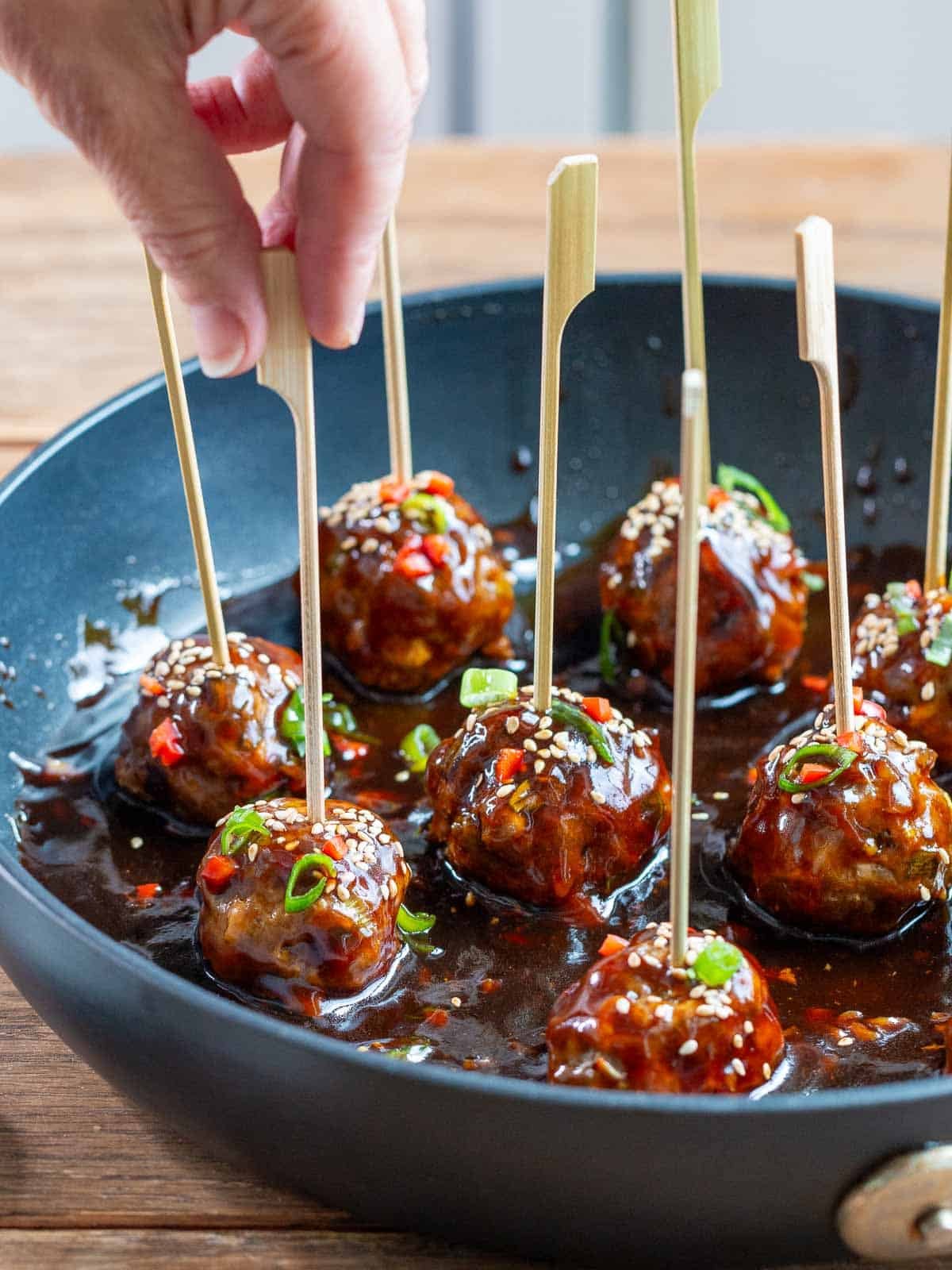 Asian Style meatballs in a skillet with bamboo skewers and a person grabbing a meatball. 