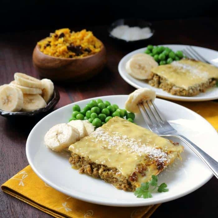 Slice of bobotie on a white plate with sliced bananas, rice with raisons and peas. 