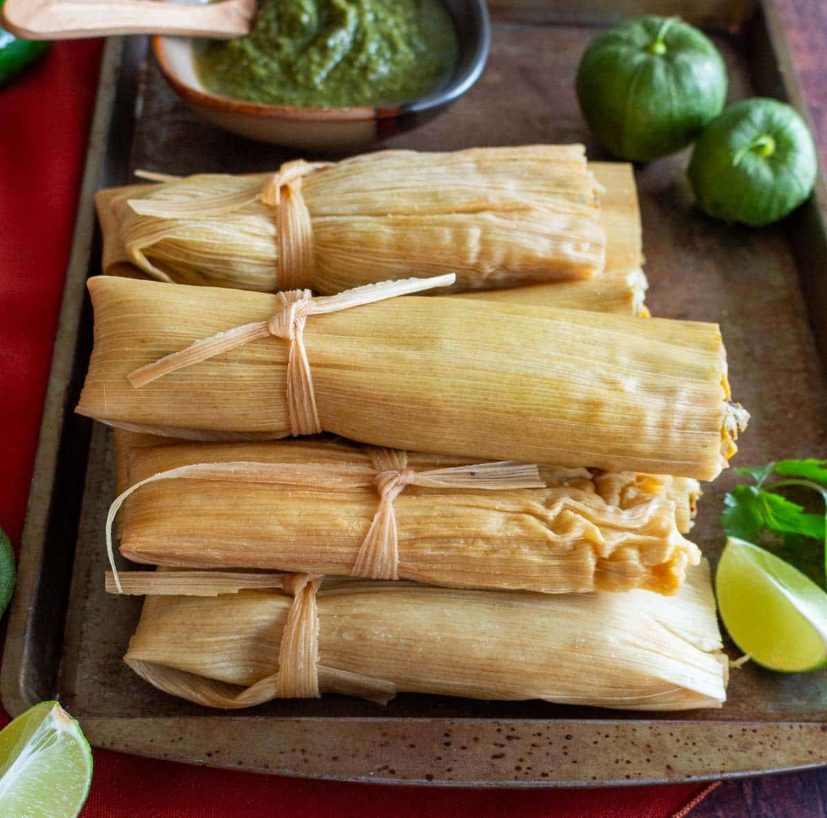 Vegetarian tamale verde on a sheet pan with a side of verde sauce and limes. 