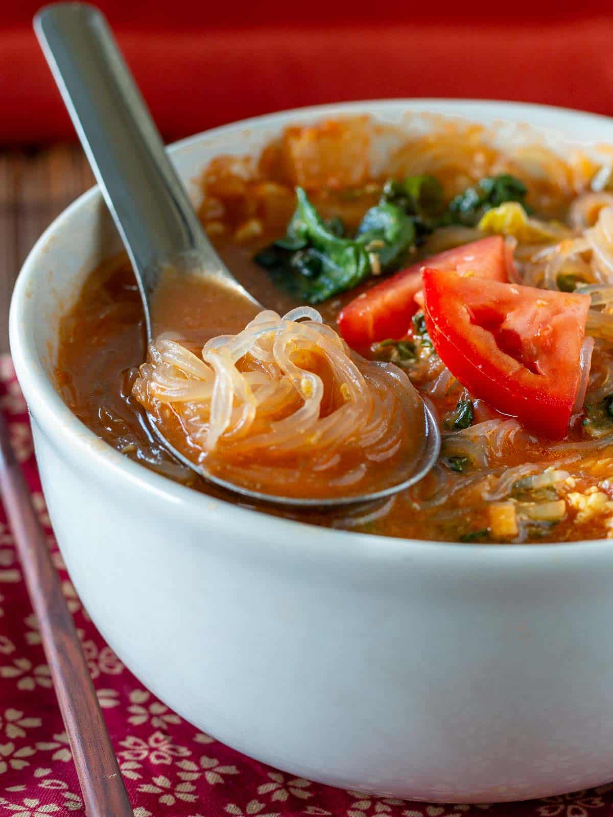 Spoonful of cooked glass noodles in a Chinese spoon nestled in a bowl of rich tomato broth. 