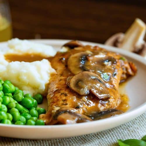 Mushroom chicken with brandy sauce on a white plate with sauce over mashed potatoes and a side of peas.