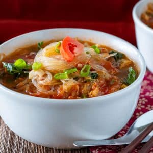 Spicy tomato glass noodle soup in a white bowl with a Chinese spoon and chopsticks.